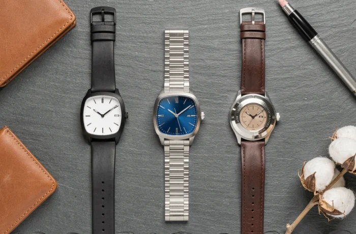 Selection of three square watches for men arranged in a flat lay on slate with leather accessories, white, blue, and black dials